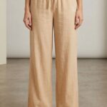 A woman models Beige wide-leg linen pajama pants, featuring an elastic waistband with drawstring and small bear embroidery, standing barefoot on a light floor.