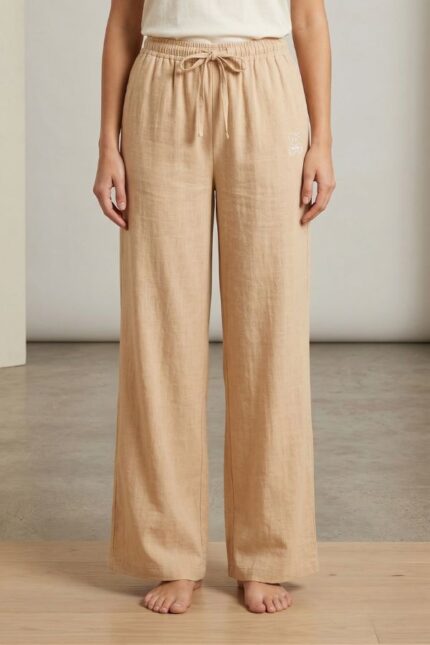 A woman models Beige wide-leg linen pajama pants, featuring an elastic waistband with drawstring and small bear embroidery, standing barefoot on a light floor.