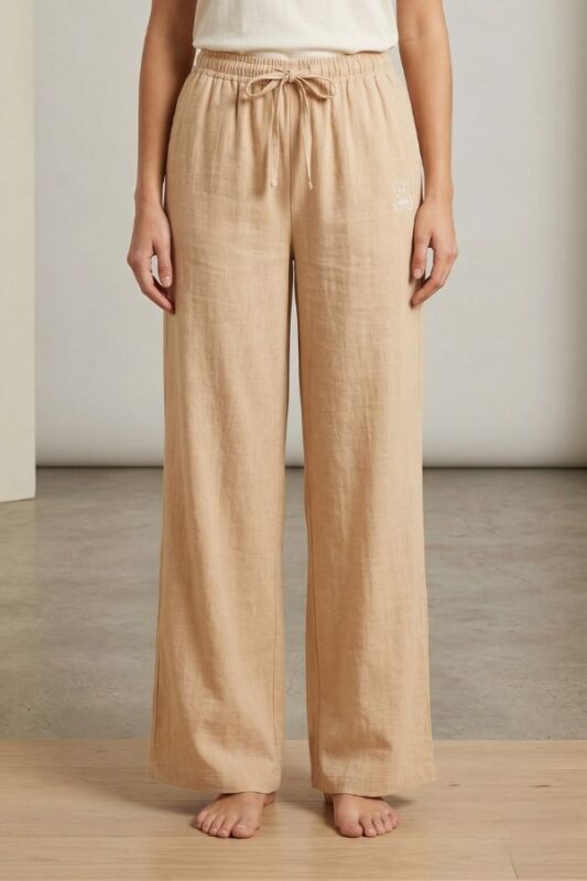 A woman models Beige wide-leg linen pajama pants, featuring an elastic waistband with drawstring and small bear embroidery, standing barefoot on a light floor.