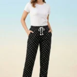 Full-body view of a model wearing comfortable black women's pajamas with white polka dots, drawstring tassels, and sneakers against a light blue background.