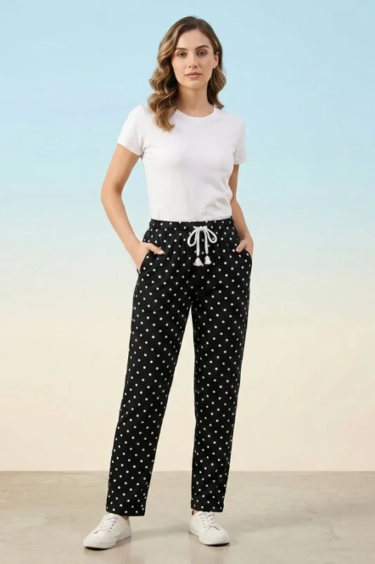 Full-body view of a model wearing comfortable black women's pajamas with white polka dots, drawstring tassels, and sneakers against a light blue background.