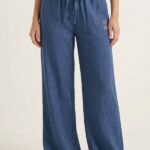 A woman stands modeling a matching indigo-blue linen top and wide-leg pajama pants, which have a tied drawstring waist and bear embroidery.