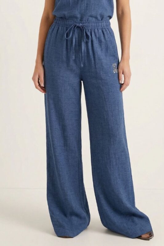 A woman stands modeling a matching indigo-blue linen top and wide-leg pajama pants, which have a tied drawstring waist and bear embroidery.