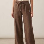 A full-body photo of a barefoot woman standing against a plain background, wearing dark brown wide-leg linen pajama pants with an elastic waist and drawstring.