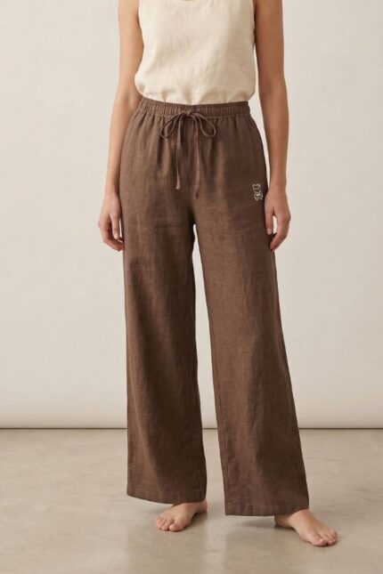A full-body photo of a barefoot woman standing against a plain background, wearing dark brown wide-leg linen pajama pants with an elastic waist and drawstring.