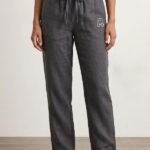 A front shot of a barefoot woman standing against a plain background, modeling charcoal gray wide-leg linen pajama pants paired with a white t-shirt.