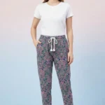 Full-body view of a model wearing elegant navy blue paisley print women's pajamas with traditional designs, drawstring with tassels, and hands in pockets against a shaded pink/blue background.