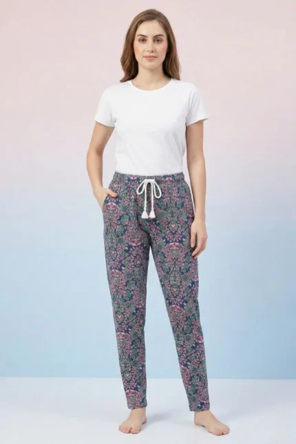 Full-body view of a model wearing elegant navy blue paisley print women's pajamas with traditional designs, drawstring with tassels, and hands in pockets against a shaded pink/blue background.