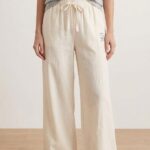 A barefoot woman stands modeling Off-White wide-leg linen pajama pants, paired with a gray t-shirt. The pants have a tied drawstring waist and bear embroidery.