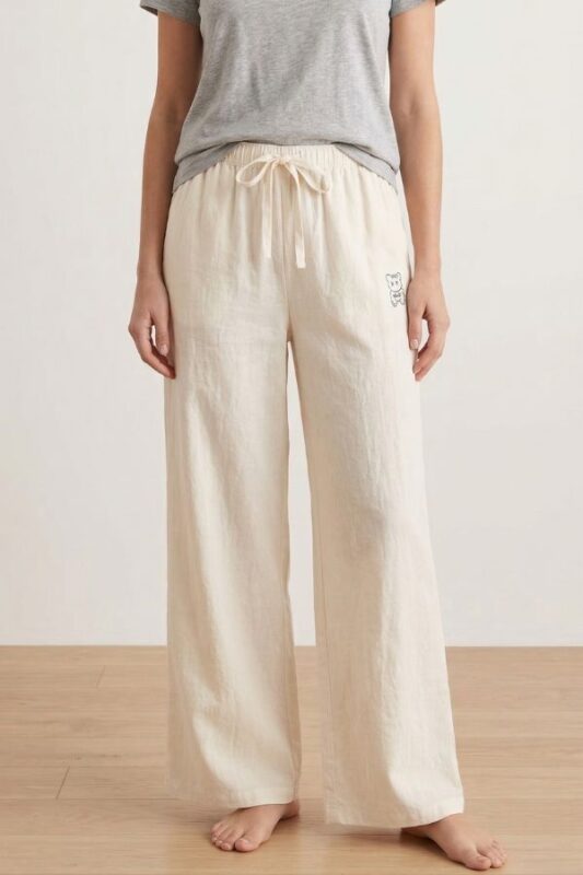A barefoot woman stands modeling Off-White wide-leg linen pajama pants, paired with a gray t-shirt. The pants have a tied drawstring waist and bear embroidery.