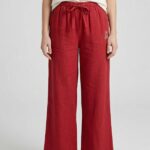 A full shot of a barefoot woman modeling Deep Red wide-leg linen pajama pants with a tied drawstring waist and a small bear logo, standing against a plain background.