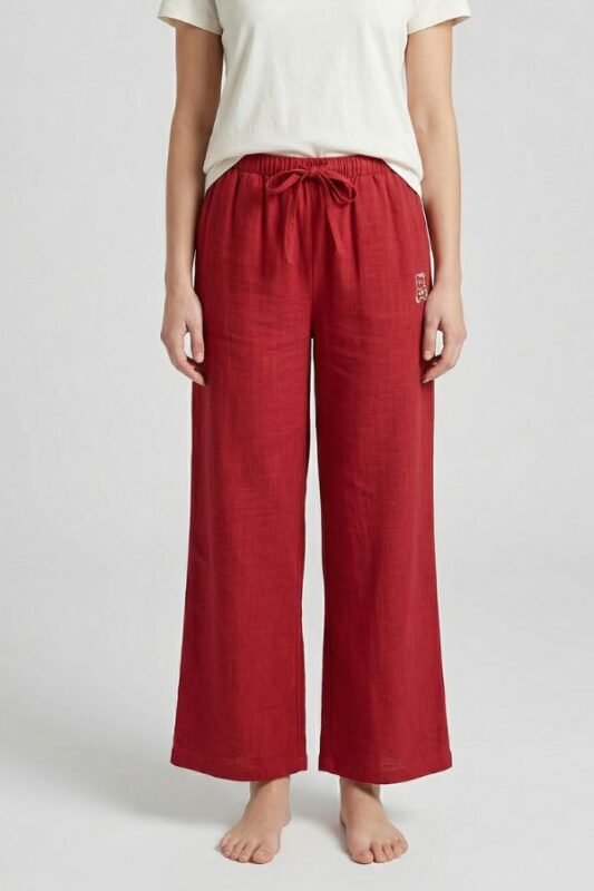 A full shot of a barefoot woman modeling Deep Red wide-leg linen pajama pants with a tied drawstring waist and a small bear logo, standing against a plain background.