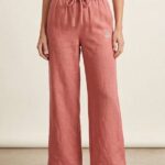 A full-length photo of a woman wearing Rose-colored wide-leg linen pajama pants with an elastic waist and drawstring, standing and wearing beige slides.